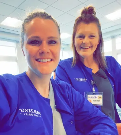 Two Herzing University students in blue scrub jackets pose for a selfie, smiling and wearing ID badges, with a bright office background.